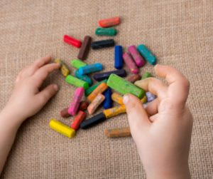 ID: Unwrapped, used crayons on a burlap tablecloth with a child's hands.